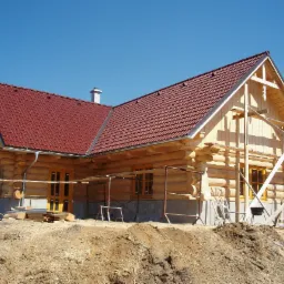 Surowy stan budowy domu z bali z czerwonym dachem, widoczne rusztowania i wykop ziemny, błękitne niebo w tle.